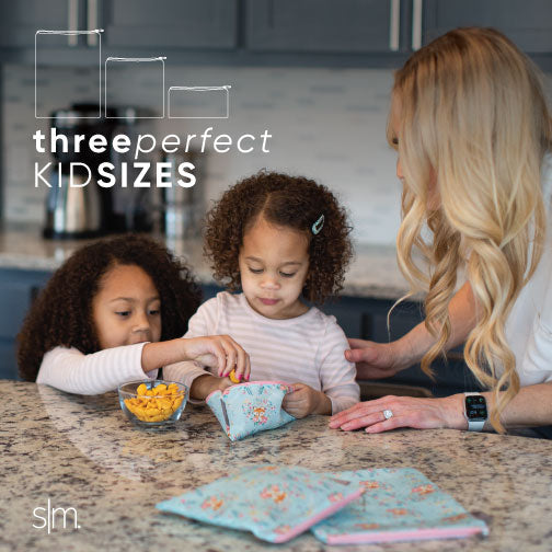 Ellie Snack Bag product displayed in a kitchen setting with neutral gray and white background, warm wood cabinetry, and soft pastel children's clothing visible.
