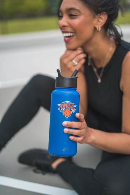 Royal blue water bottle held by person wearing black sleeveless top with green grass and neutral background