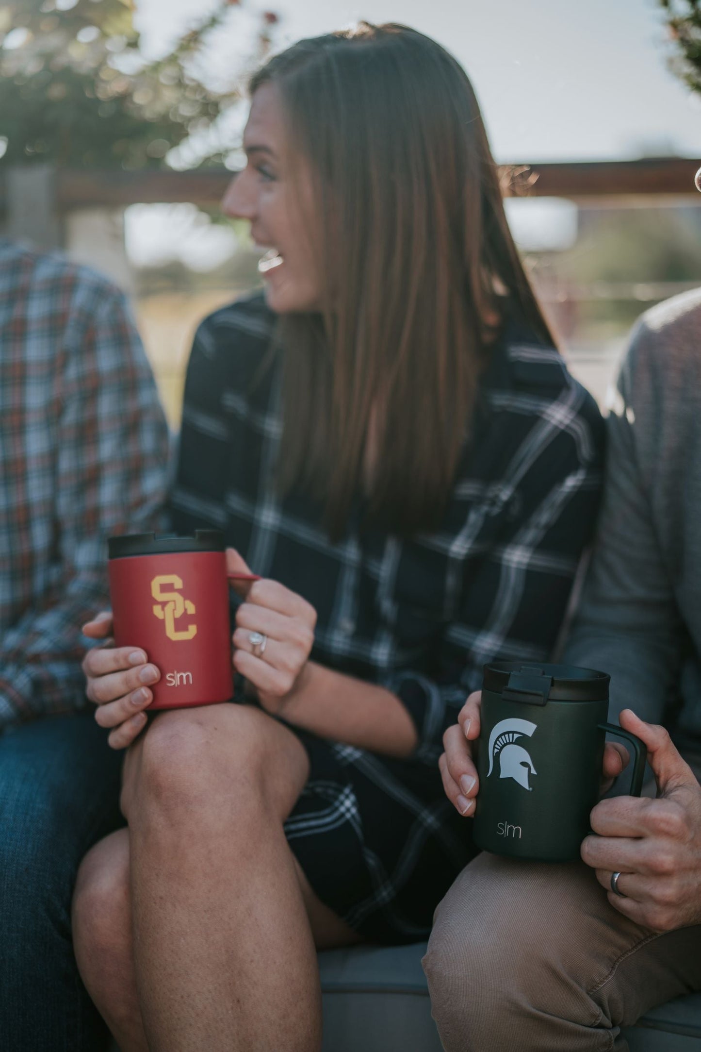 Woman in center holding bright red tumbler with gold accents, person beside her with dark forest green tumbler, warm natural tones throughout