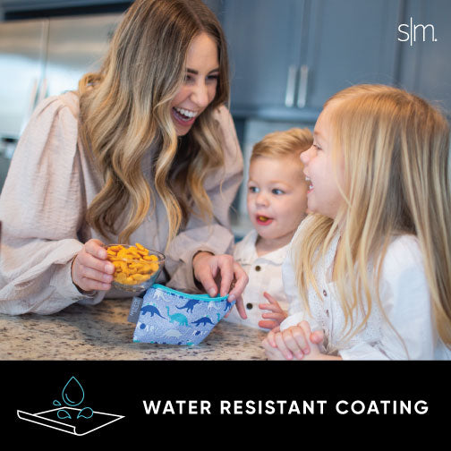 Woman in beige sweater holding yellow-orange snack bag in front of warm gray kitchen cabinets, demonstrating product packaging and size.