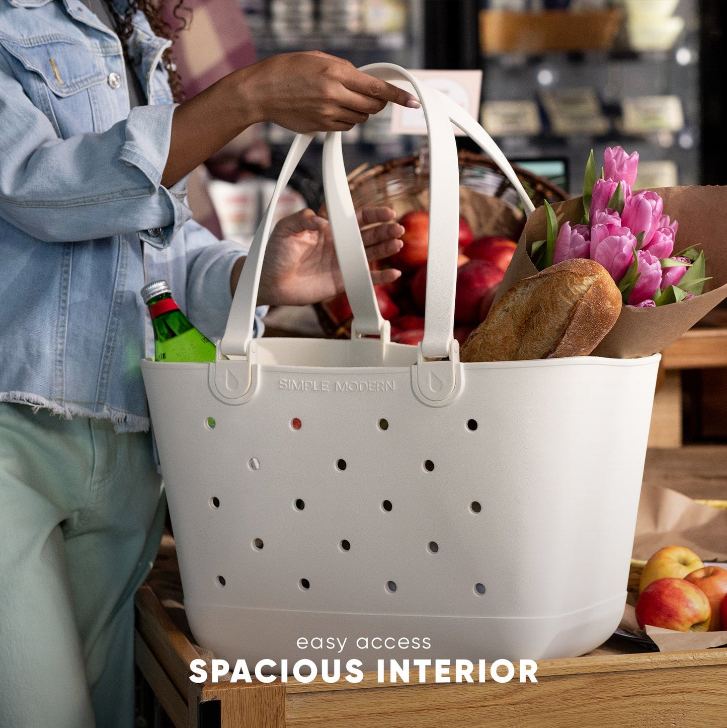 White Simple Modern tote bag with perforated design held by person wearing chambray shirt, filled with pink tulips, bread, apples, and green bottle. Wooden table with fresh produce and flowers visible.