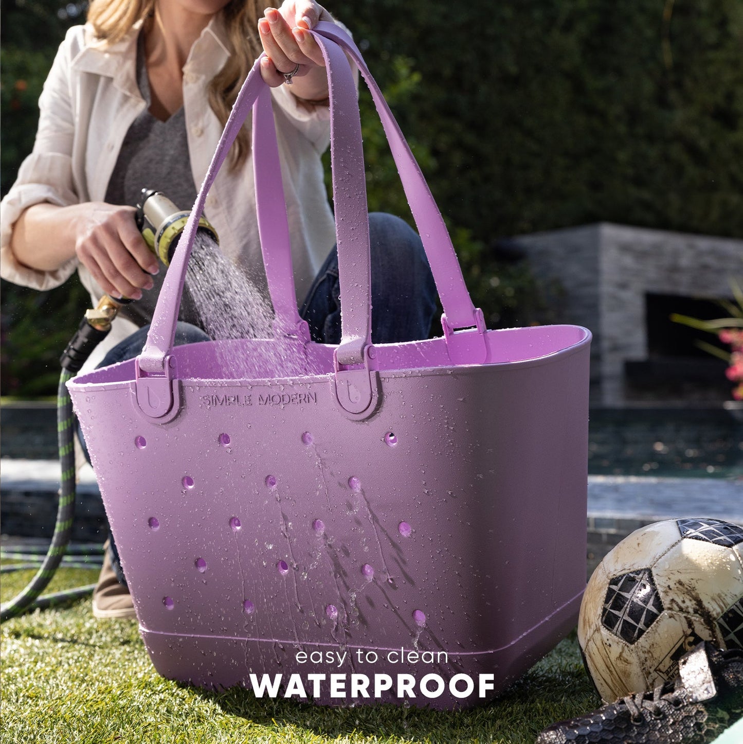 Woman demonstrating waterproof Simple Modern Getaway Bag in lavender-purple while spraying it with a garden hose. Bag features sturdy handles and perforated design. Text overlay reads "easy to clean" and "WATERPROOF."