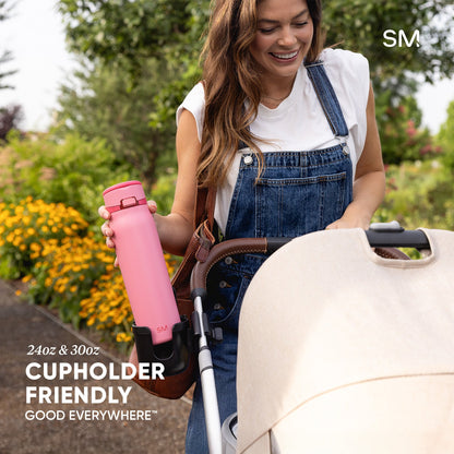 Woman holding pink Simple Modern water bottle while pushing stroller in sunny garden. Bottle features cupholder-friendly design in 24oz and 30oz sizes. Text reads "Good Everywhere."