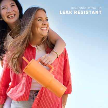 Two women smiling outdoors wearing red pinstriped blazers, holding a bright orange leak-resistant insulated tumbler with straw lid against a blue sky background.